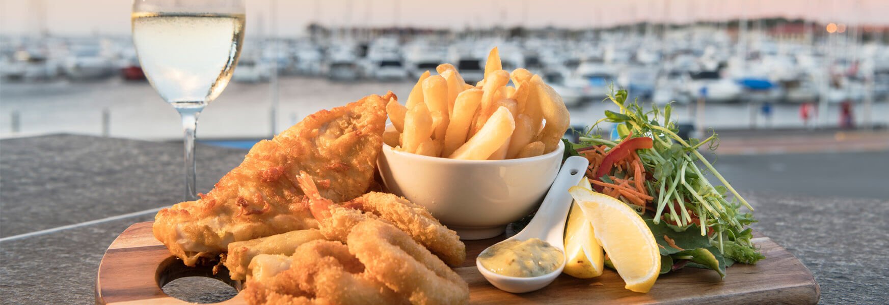 A fish and chips dish on a wooden board besides a glass of white wine, with the Hillary's boat harbour in the background